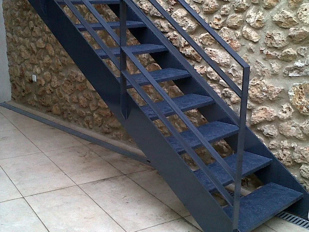 Escalera de metal azul contra una pared de piedra en una zona interior con piso de baldosas.