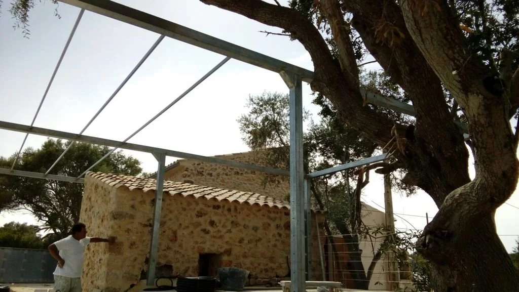 Una estructura de metal en construcción junto a una antigua casa de piedra y un árbol grande. Hay una persona de pie observando la estructura.
