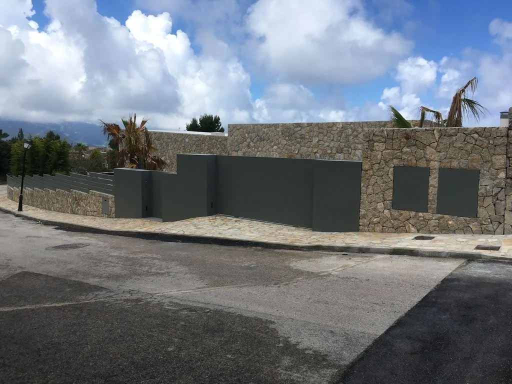 Muros de piedra y una reja gris en un área urbana con árboles y un cielo nublado.