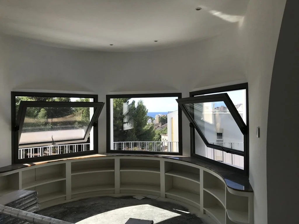 Ventanas abiertas con vista a árboles y edificios en una habitación en construcción, con estanterías blancas curvas debajo de las ventanas.