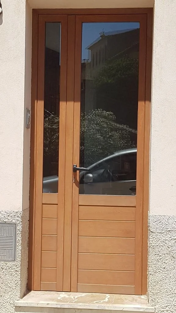 Puerta de madera con marco de cristal transparente y paneles inferiores de madera, ubicada en una entrada con muro pintado de blanco y base de piedra.