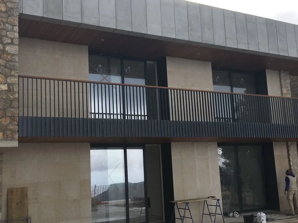 Edificio en construcción con paredes de piedra y paredes de cemento, balcón con barandal de barras verticales y ventanas de cristal.