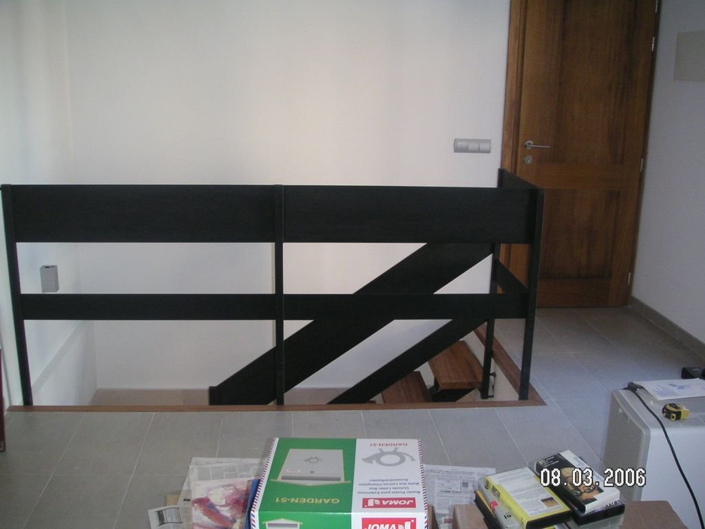 Vista de un cuarto con escaleras de madera y barandal negro, puerta de madera y papel y herramientas sobre la mesa.