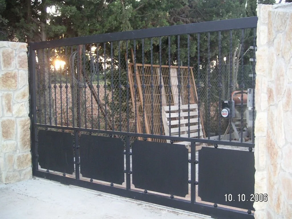 Puerta de rejas de metal negro con pilares de piedra en los lados, en el fondo se ven maderas y una cortina de la cerca, con árboles y el sol al atardecer.