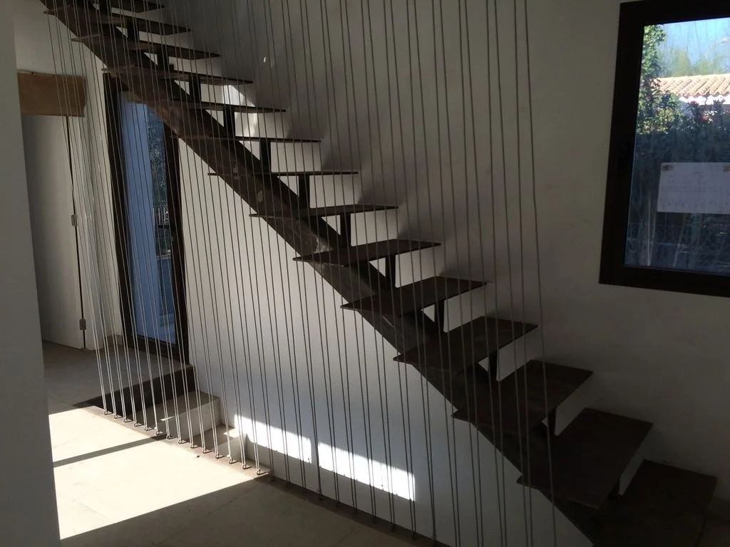 Escalera moderna con barandales de cables metálicos y un ventanal grande a la derecha, en un interior con paredes blancas y piso claro.