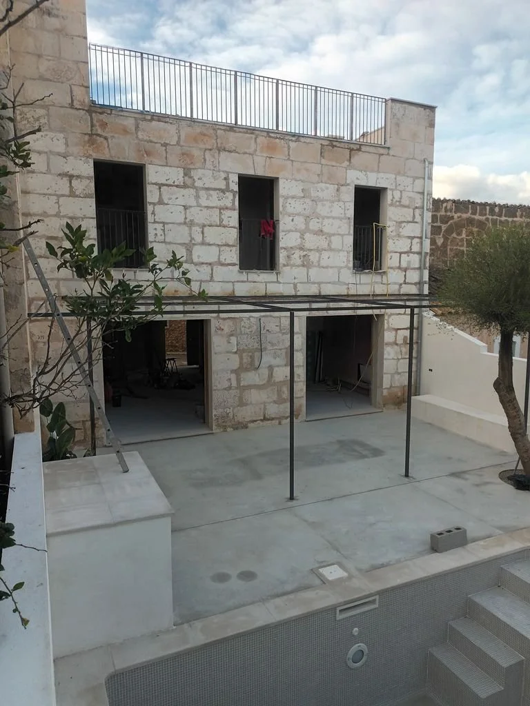 Construcción de una casa de dos pisos con paredes de bloques y ventanas abiertas, en un patio con una piscina vacía, árboles y un cielo parcialmente nublado.