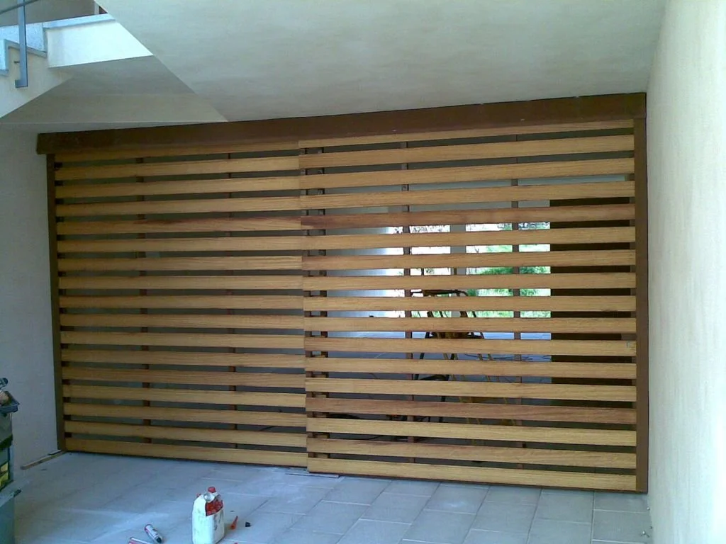 Puerta corrediza de madera con listones horizontales, parcialmente abierta, en un área exterior.