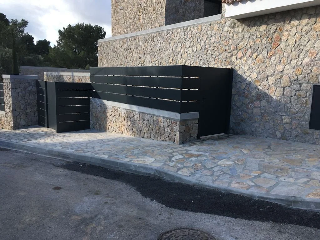 Cerca moderna de metal negro en una entrada de piedra en un muro de piedra natural, con una acera de piedra y una calle de pavimento.
