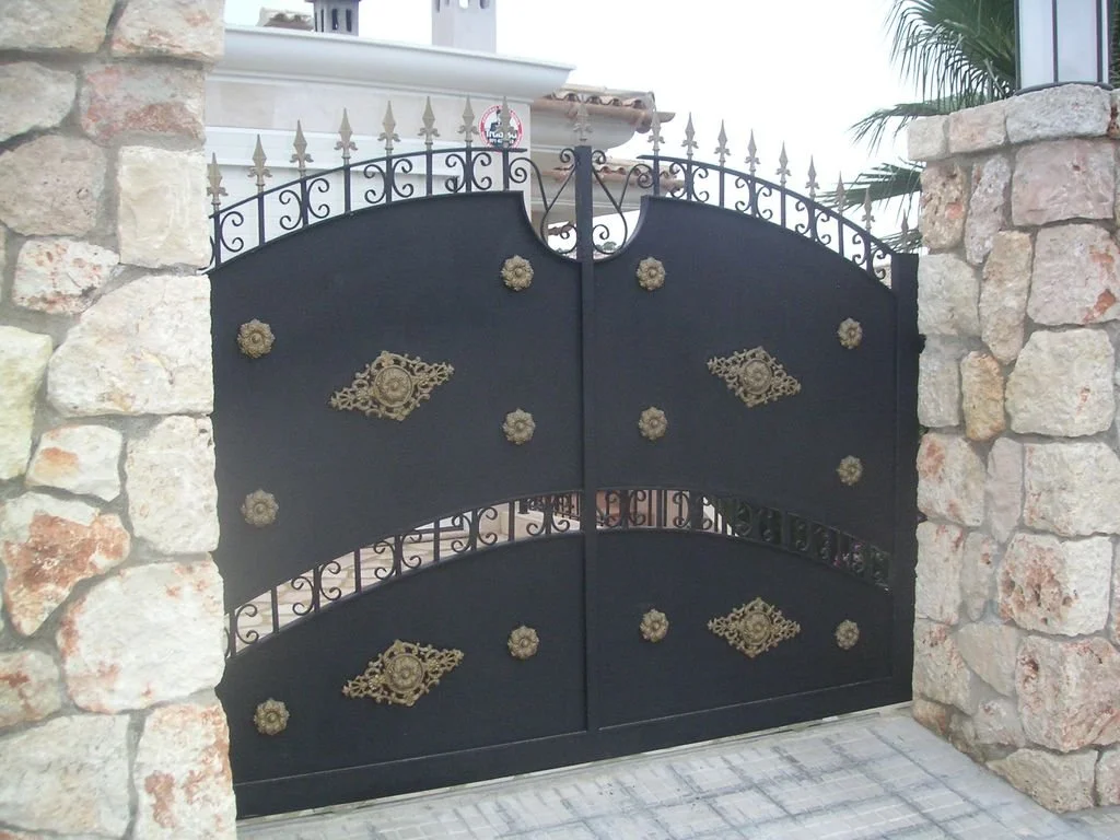 Una puerta de reja negra con adornos dorados, rodeada de muros de piedra.