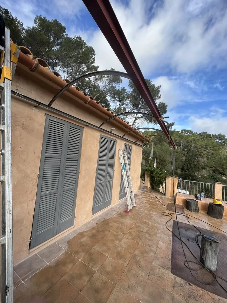 Imagen de una terraza exterior junto a una casa con ventanas de celosía, un andamio y una escalera, con cables y herramientas, durante el día con cielo parcialmente nublado.