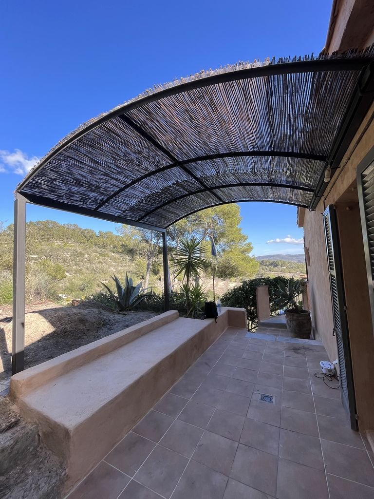 Terraza exterior con suelo de baldosas, bancos de cemento, plantas de cactus y árboles de fondo, con un toldo de cañas de bambú y cielo azul claro.