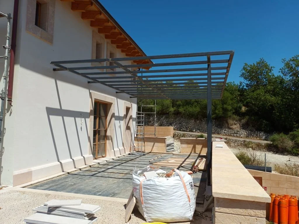 Estructura metálica en construcción en un patio exterior, con una casa de fondo y árboles en la distancia, cielo despejado.
