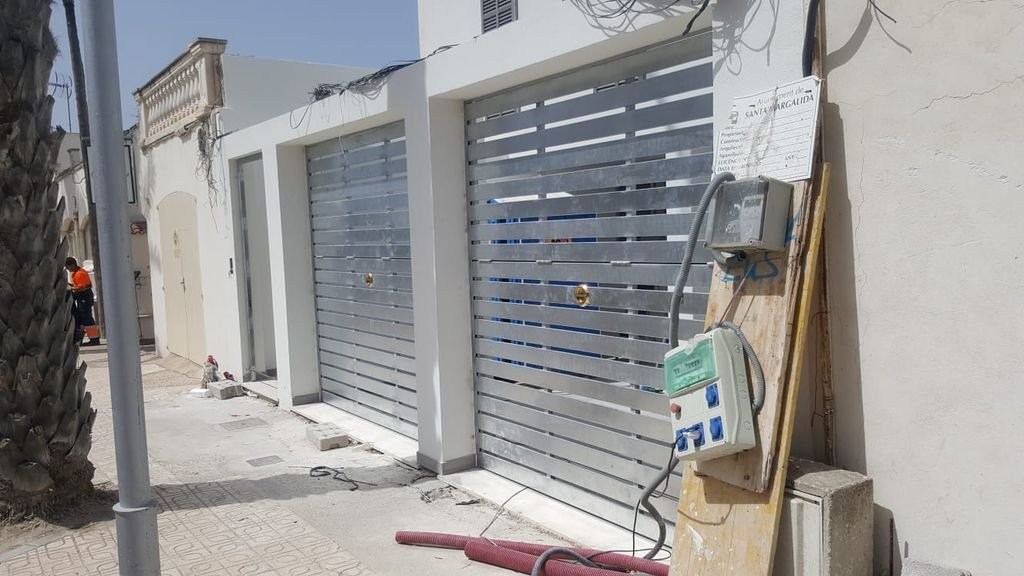 Puertas metálicas en una vivienda, en la calle hay una palmera y una falta de cableado en la pared.
