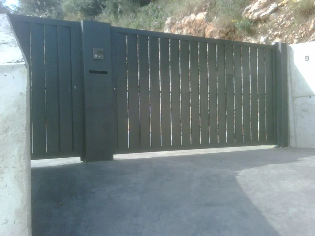 Puerta de metal pintada de negro con un buzón incorporado y instalada entre dos muros de concreto, en una entrada posiblemente de una propiedad privada.