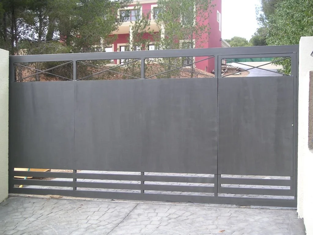 Puerta de metal gris en un muro de entrada a una propiedad, con un edificio rosa y árboles en el fondo.