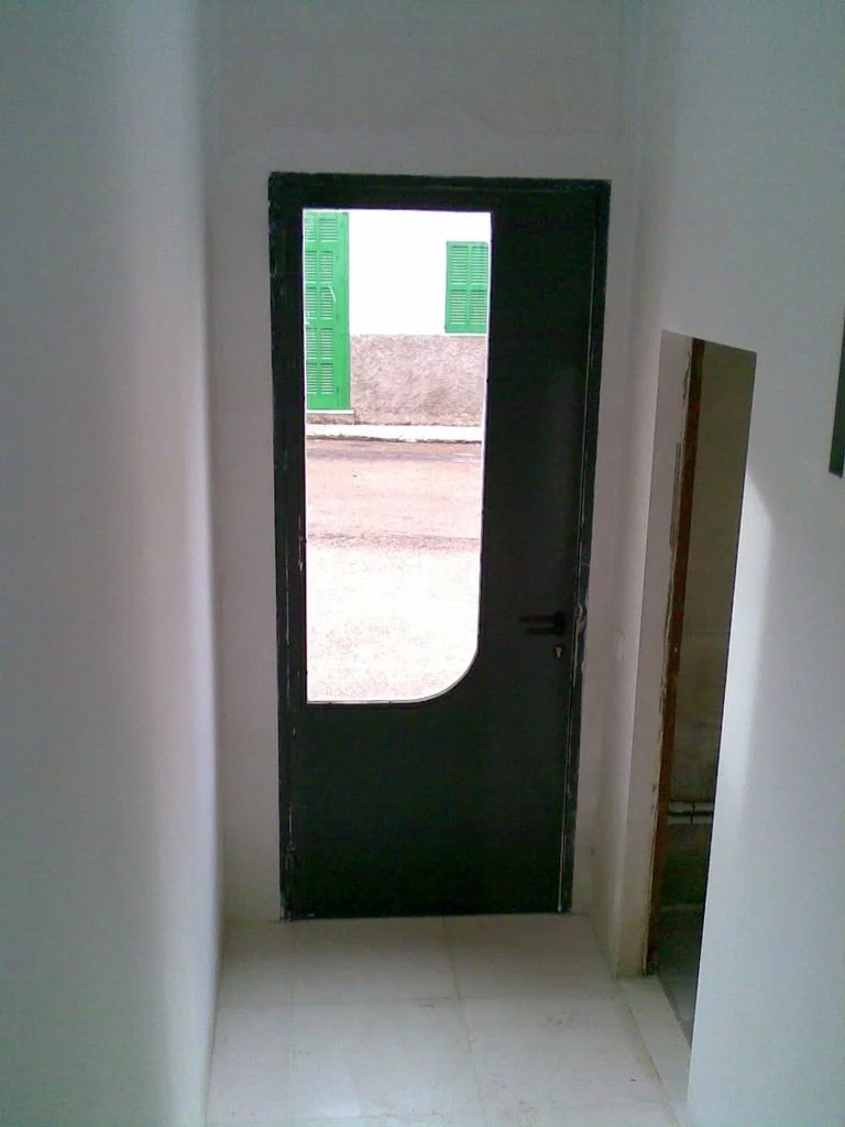 Puerta negra de metal con ventana de vidrio, vista desde el interior de una casa, con paredes blancas y piso de azulejos blancos.