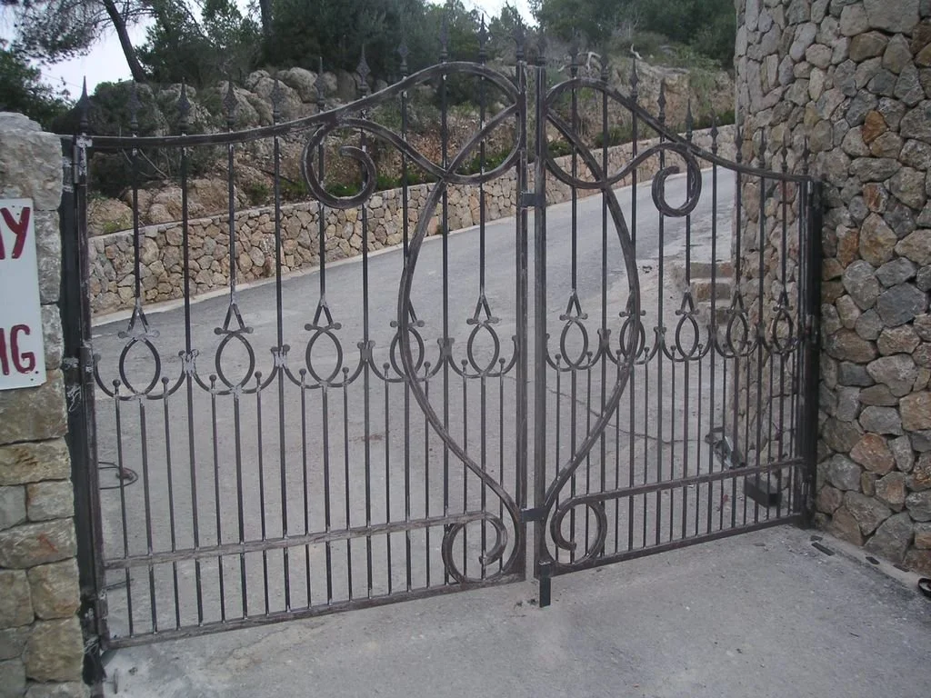 Una puerta de reja de metal con diseño de corazones y bucles, ubicada entre muros de piedra, con una calle de fondo.