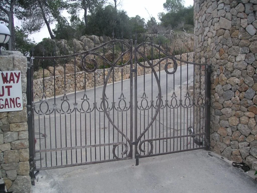 Puerta de reja de hierro con decoración de corazón, entre muros de piedra.