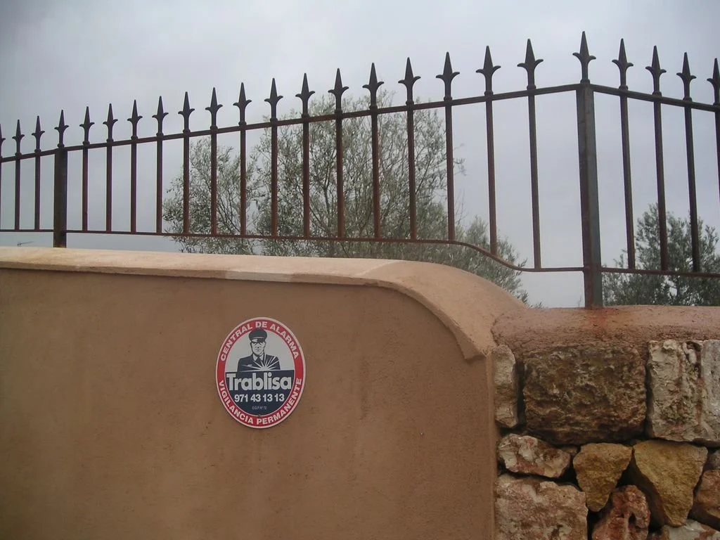 Valla de metal negro con picos sobre muro de piedra, cartel con logo y texto "Trabaja" y número de contacto, cielo nublado y árboles en el fondo.