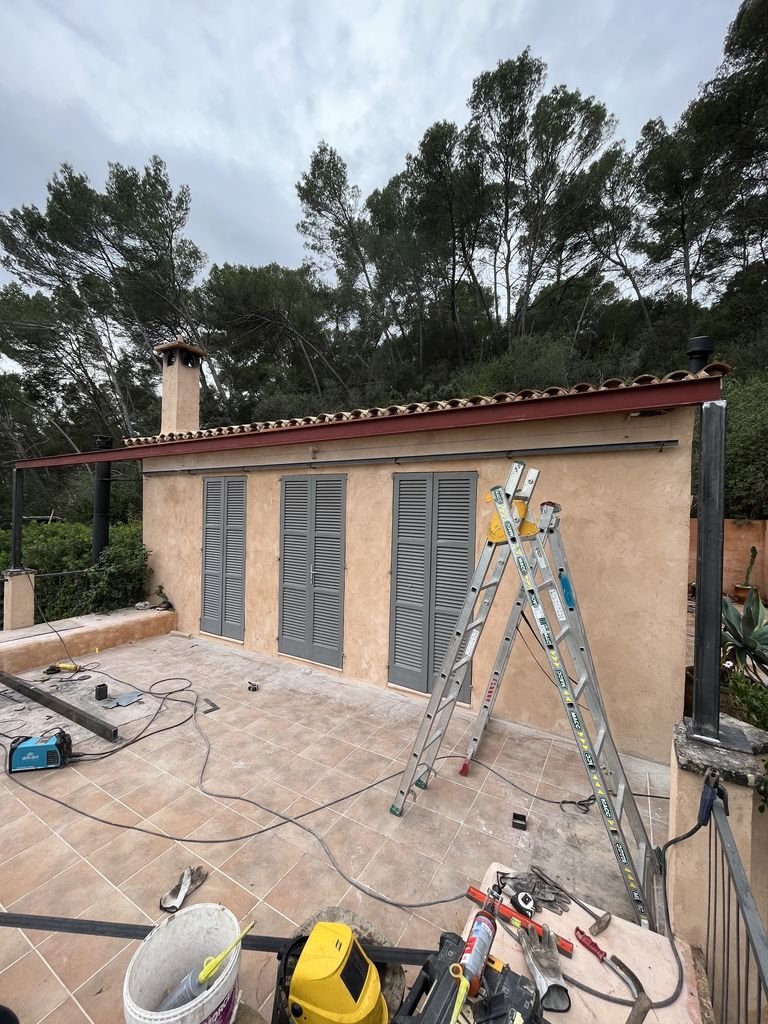 Casa en construcción con ventanas con persianas cerradas, en un patio con herramientas y escaleras, rodeada de árboles.