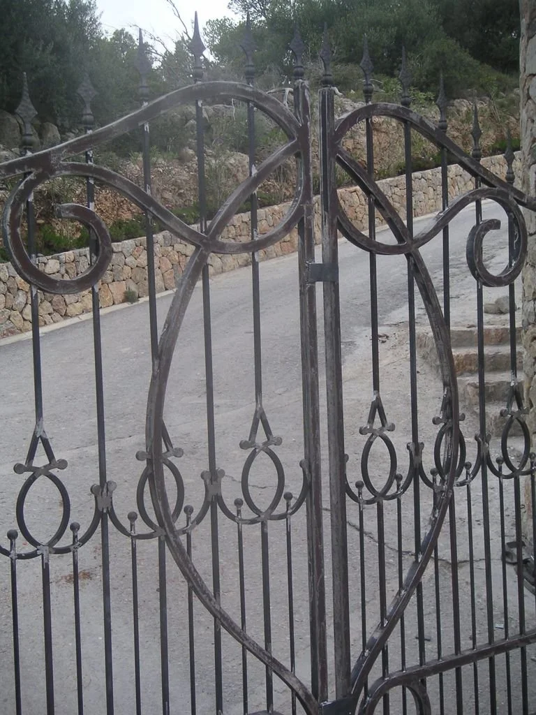 Barandilla de metal con forma de corazón y detalles decorativos. Visto desde cerca con una calle y un muro de piedras en el fondo.