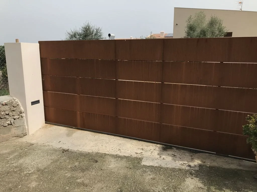 Muro de cerca de madera en color marrón con una estructura blanca al lado, en un ambiente exterior con suelo de concreto y vegetación en el fondo.