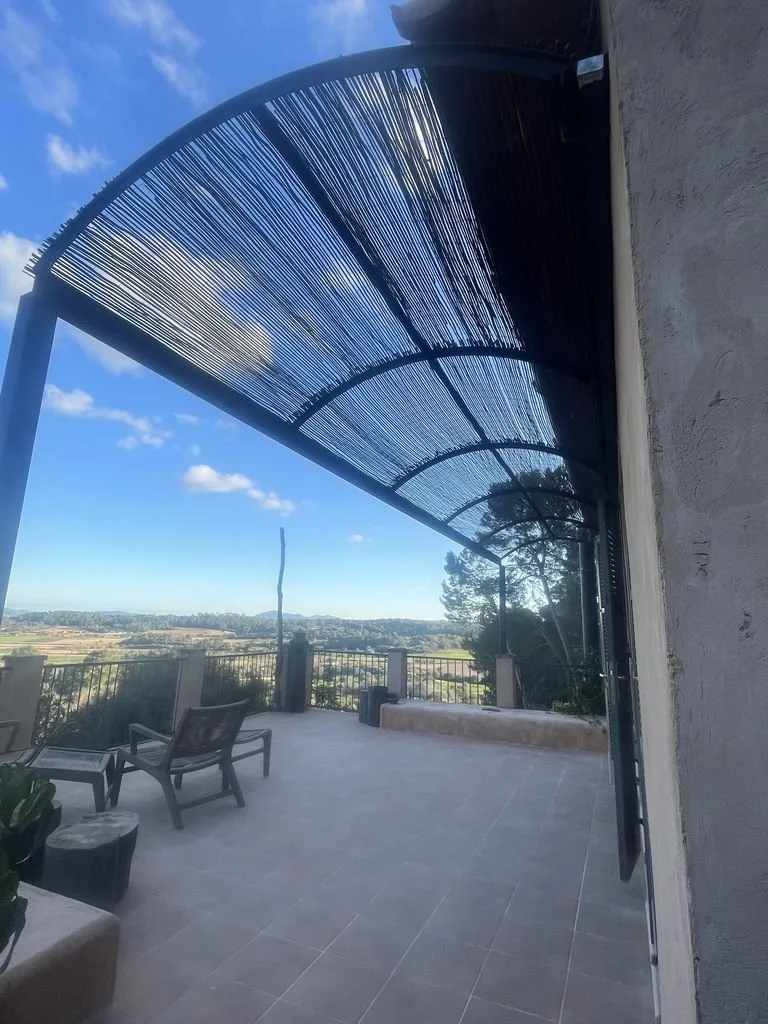 Vista de una terraza con sillones y una estructura de sombra de ramas, con un paisaje de campo y árboles al fondo bajo un cielo azul con nubes.