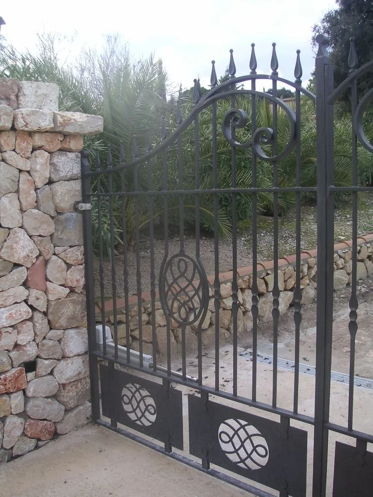 Reja de hierro negro con diseño decorativo, muro de piedras a la izquierda y plantas verdes en el fondo.