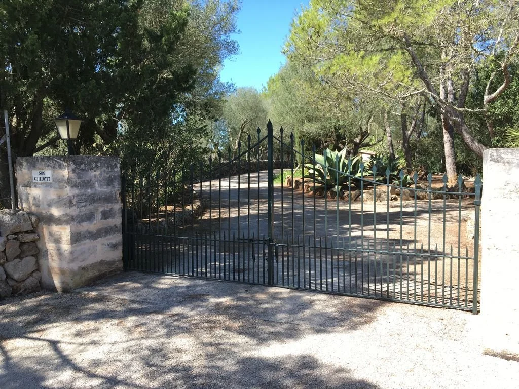 Entrada de un camino de tierra con puerta de rejas negras, rodeada de árboles y plantas en un entorno natural