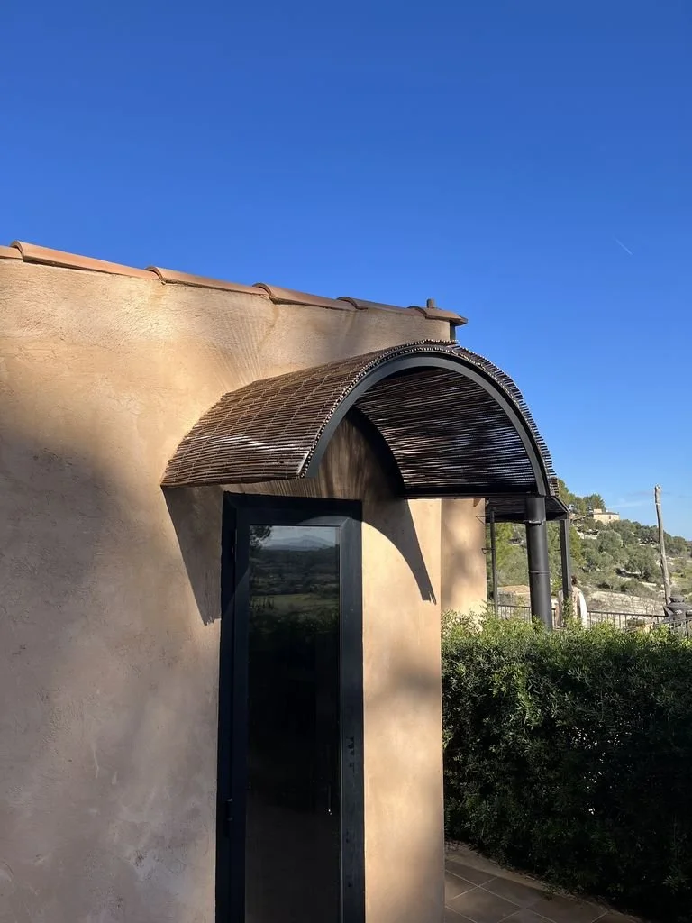 Puerta de metal con estructura de sombra de mimbre en forma de arco en una pared beige, con cielo azul y vegetación en el fondo.