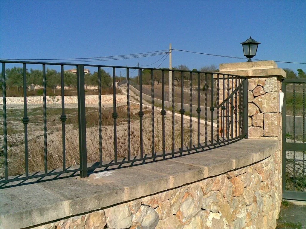Reja de metal en piedra que rodea una entrada a una carretera o camino en un área rural.