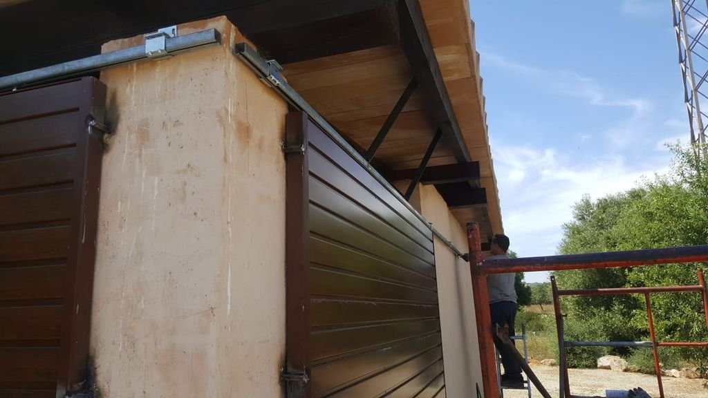 Trabajador instalando una lámina de metal en el cerramiento de un edificio en construcción, con andamios y vegetación a un lado.