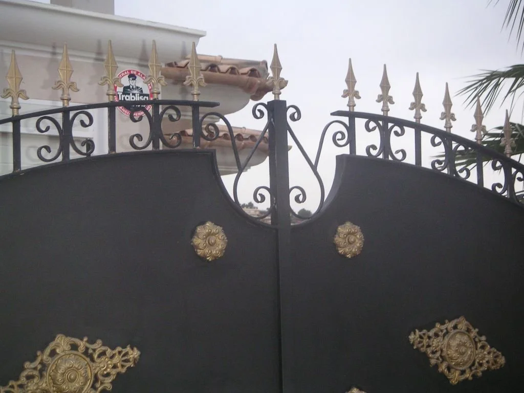 Puerta negra con detalles dorados y remates en forma de flecha en la parte superior, con una cerca de varillas de hierro decoradas y un letrero en la pared del fondo."}