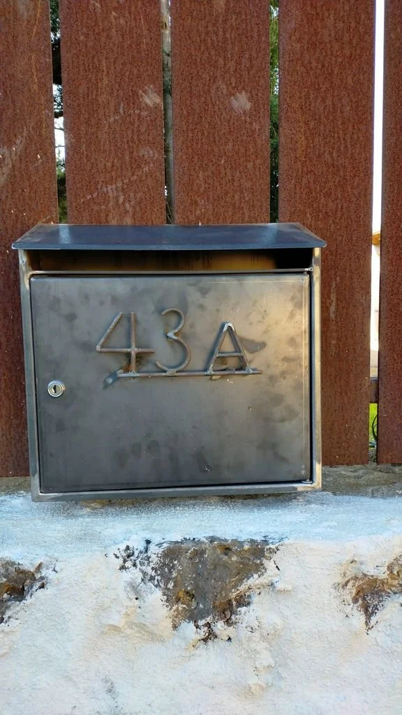 Caja de correo de metal con el número 43A, instalada en una pared con listones de madera y una base de concreto con desgaste.