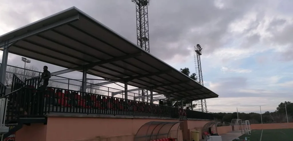 Gradas de un campo de fútbol con un área techada y una persona. Líneas de iluminación y porterías en el campo.