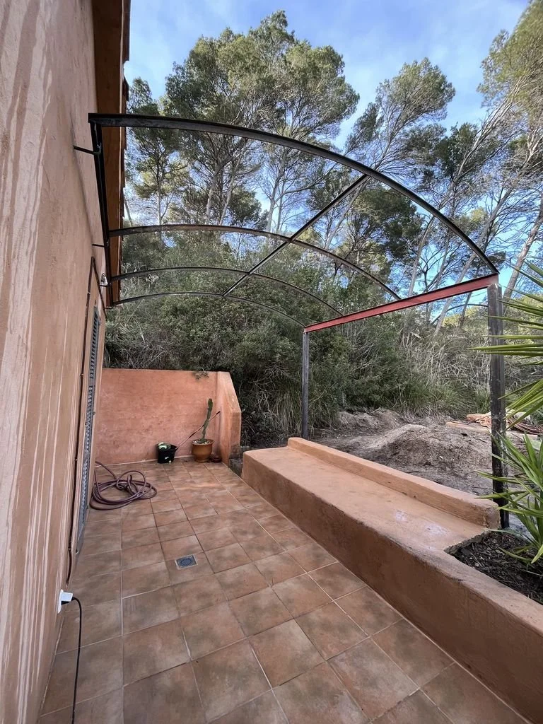 Un balcón con paredes de color terracota, tejas de piso, con un par de macetas con cactus, manguera sin usar, y un techo de arcos de metal y lámina traslúcida, rodeado de árboles y naturaleza.