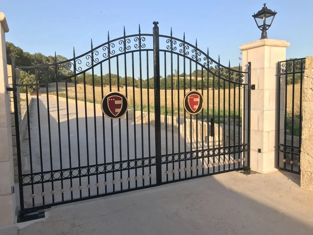 Puerta metálica negra con decoración de círculos y pinchos en la parte superior, flanqueada por un poste de piedra con farol, durante el día con cielo despejado y campo de campo en el fondo.