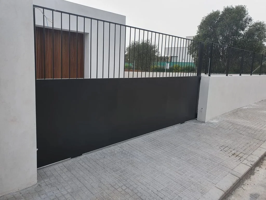 Valla de hierro y muro de concreto en un entorno urbano con árboles al fondo.
