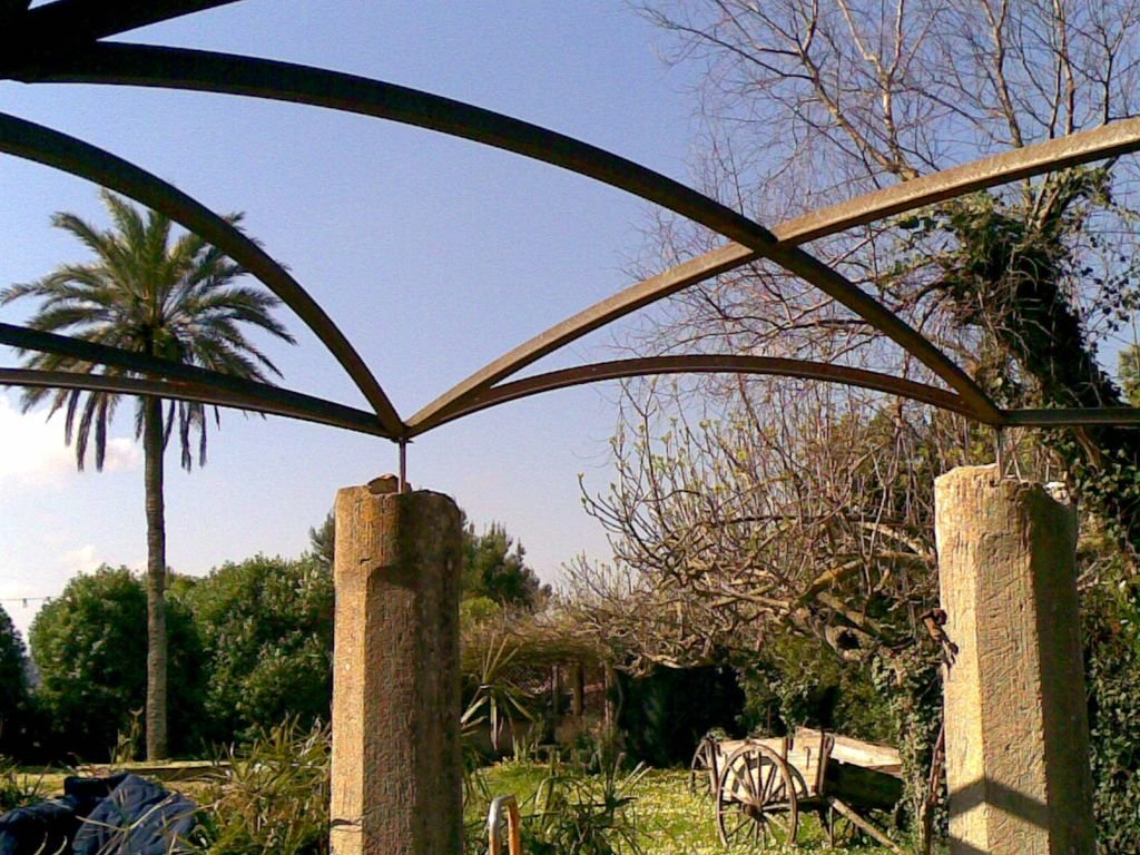Estructura metálica en forma de medio círculo en un jardín con árboles y un carruaje de madera.