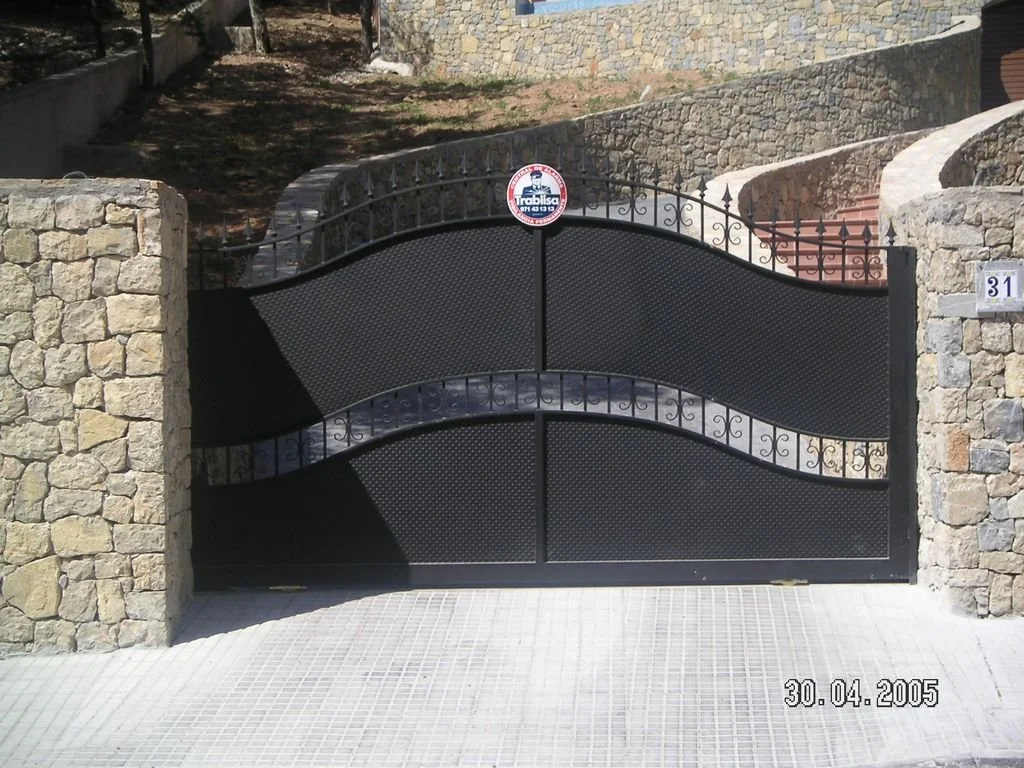 Reja negra de metal en una entrada de casa, rodeada por muros de piedra, con señal de advertencia y número 31.
