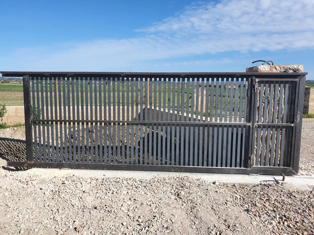 Reja negra de metal en un suelo de piedra, con un fondo de campo y cielo azul con nubes.