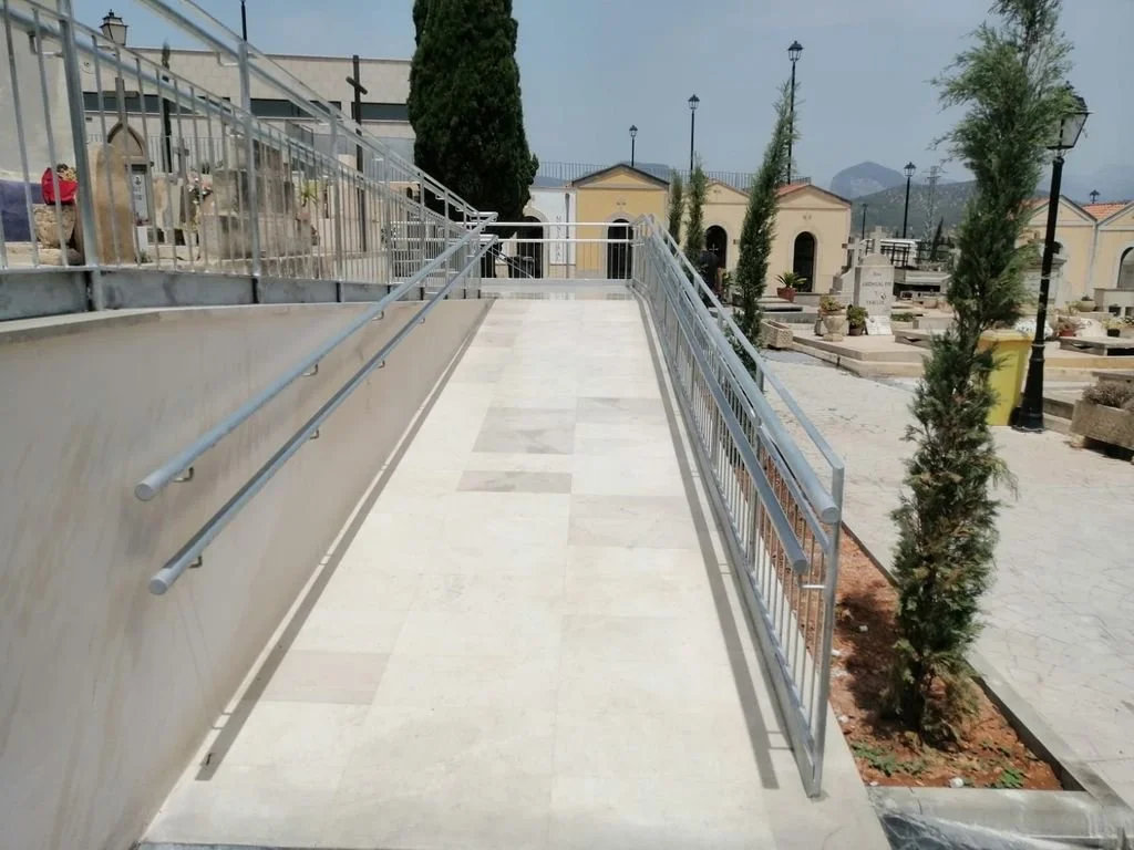 Un paseo elevado con barandales de metal y rampas en un cementerio con lápidas, árboles y lámparas en el fondo