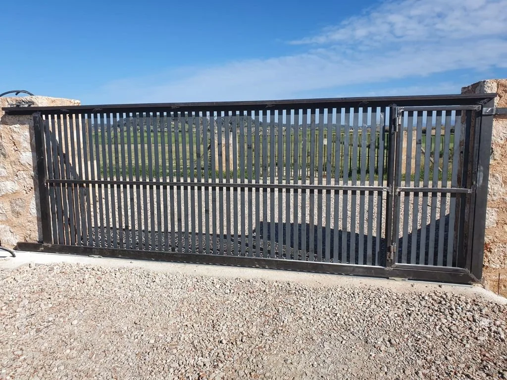 Valla de metal negro con puertas corredizas, en un entorno exterior con cielo azul y campo verde al fondo.