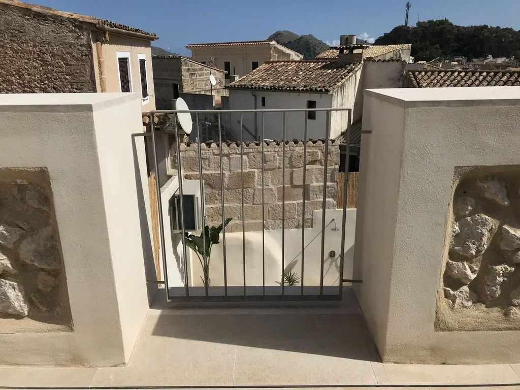 Vista desde una terraza con una verja de metal y paredes de piedra y concreto, mostrando edificios con techos de tejas y un paisaje montañoso en el fondo.