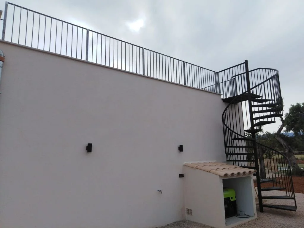 Escalera de caracol negra en un muro blanco, en un espacio exterior con cielo nublado y árboles en el fondo.