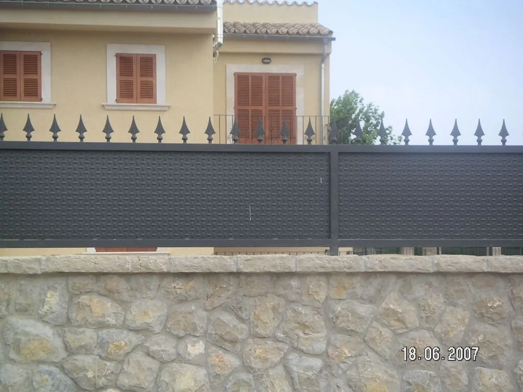 Casa con ventanas con contraventanas de madera y una cerca negra de hierro en primer plano, sobre un muro de piedra.
