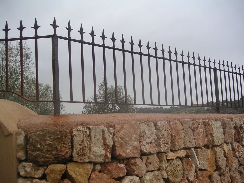 Valla de hierro con picos, sobre muro de piedra y árboles al fondo.