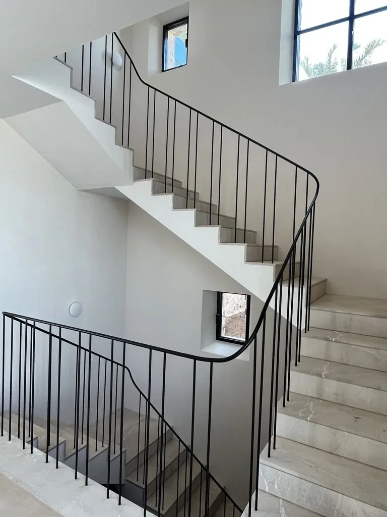 Escalera de interior moderna con barandal de metal negro, paredes blancas y ventanas pequeñas con vista al exterior.