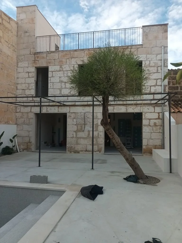 Casa en construcción con muro de piedra, un árbol inclinado, estructura de metal para alguna cobertura, en un patio con piscina en construcción y cielo nublado.