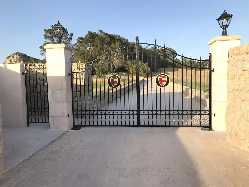 Puerta negra de reja con dos postes con faroles y distintivos en medio, que da hacia un camino con árboles al fondo y un cielo despejado.